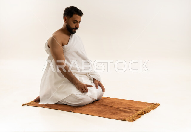الخشوع في الصلاة والتضرع إلى الله، الجلوس للتشهد على السجادة، بورتريه لرجل عربي خليجي عماني يرتدي ملابس الإحرام ويؤدي فريضة الصلاة، أداء مناسك الحج والعمرة، التقرب إلى الله بالعبادة والطاعة، خلفية بيضاء