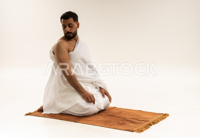 التسليم وإنهاء الصلاة، الخشوع والتضرع إلى الله، بورتريه لرجل عربي خليجي عماني يرتدي ملابس الإحرام يجلس على السجادة ويؤدي فريضة الصلاة، أداء مناسك الحج والعمرة، التقرب إلى الله بالعبادة والطاعة، خلفية بيضاء