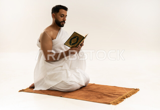 قراءة القرآن الكريم بتمعن وتركيز, تلاوة كتاب الله، بورتريه لرجل عربي عماني خليجي يرتدي ملابس الإحرام يجلس على سجادة الصلاة ويمسك بين يديه المصحف الشريف، أداء مناسك الحج والعمرة، التقرب الى الله بأداء العبادة والطاعة, خلفية بيضاء