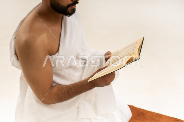 تلاوة كتاب الله، قراءة القرآن الكريم بتمعن وتركيز, بورتريه مقرب لرجل عربي عماني خليجي يرتدي ملابس الإحرام يجلس على سجادة الصلاة ويمسك بين يديه المصحف الشريف، أداء مناسك الحج والعمرة، التقرب الى الله بأداء العبادة والطاعة, خلفية بيضاء