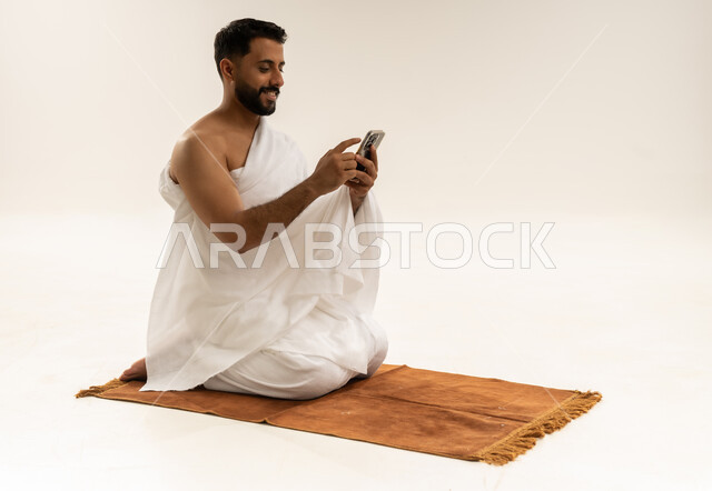 الدليل الالكتروني للحاج والمعتمر، استخدام تطبيقات الهاتف المحمول، بورتريه لرجل عربي خليجي عماني يرتدي ملابس الإحرام ويجلس على سجادة الصلاة ويمسك جهاز الجوال بيده بإيماءات السعادة، أداء فريضة الحج والعمرة، خلفية بيضاء