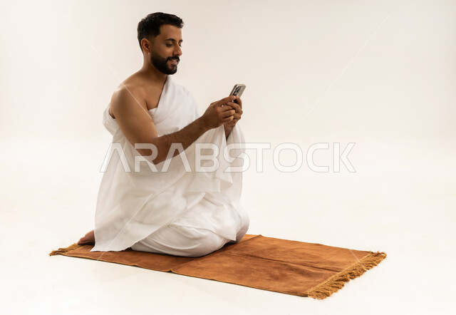 استخدام تطبيقات الهاتف المحمول، بورتريه لرجل عربي خليجي عماني يرتدي ملابس الإحرام ويجلس على سجادة الصلاة ويمسك جهاز الجوال بيده بإيماءات السعادة، الدليل الالكتروني للحاج والمعتمر، أداء فريضة الحج والعمرة، خلفية بيضاء