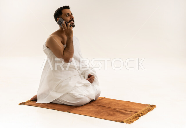إجراء مكالمة هاتفية باستخدام الهاتف المحمول، بورتريه لرجل عربي خليجي عماني يرتدي ملابس الإحرام يجلس على سجادة الصلاة ويتحدث عبر الجوال، استخدام جهاز تقني حديث ومتطور، أداء فريضة الحج والعمرة، خلفية بيضاء