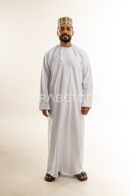 الوقوف باستقامة بتعابير الثقة بالنفس، بورتريه لشاب عربي خليجي عماني مبتسم يرتدي الدشداشة والكمه ينظر إلى الكاميرا بايماءات السرور والبشاشة، الأناقة والاهتمام بالمظهر الخارجي، الجسم بالطول الكامل، خلفية بيضاء