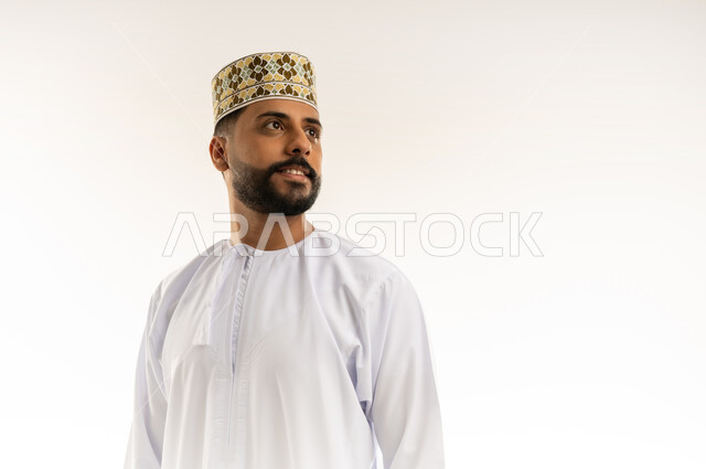 Gestures of distraction and deep thought, contemplation and future planning, close-up portrait of a smiling young Omani Gulf Arab man wearing a dishdasha and a sleeve standing and looking somewhere, elegance and interest in outward appearance, white background