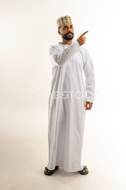 تحديد موقع شيء ما، الإشارة باصبع السبابة نحو مكان ما محدد، بورتريه لشاب عربي خليجي عماني مبتسم يرتدي الدشداشة والكمه ينظر بعيدا بايماءات الشرود، الوقوف بثقة واستقامة، الاهتمام بالمظهر الخارجي، الجسم بالطول الكامل، خلفية بيضاء