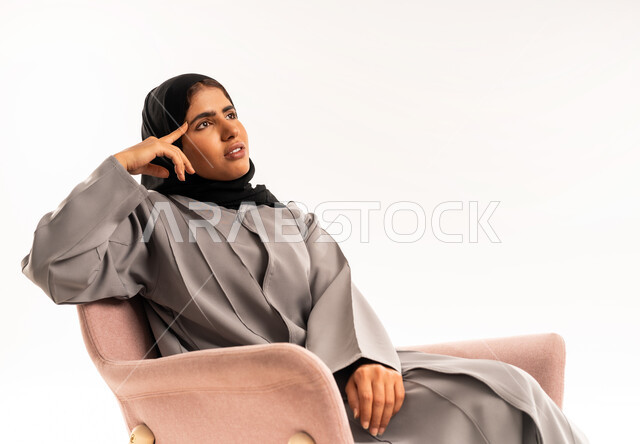 مواجهة المشكلات والتحديات في الحياة العملية، التفكير لإيجاد حل مشكلة ما، شرود الذهن والتأمل والتخطيط للمستقبل، بورتريه لامرأة عربيه خليجيه سعوديه محجبه ترتدي العباءة تجلس على كرسي مريح وتنظر الى الاعلى بإيماءات التركيز، خلفية بيضاء