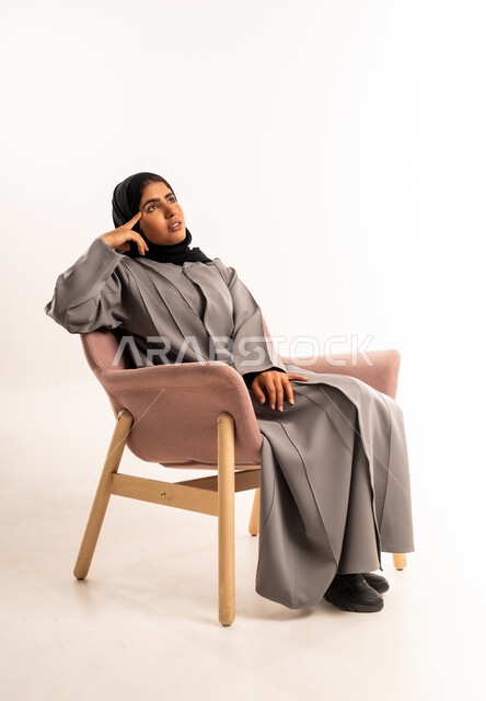 التفكير لإيجاد حل مشكلة ما، شرود الذهن والتأمل والتخطيط للمستقبل، بورتريه لامرأة عربيه خليجيه سعوديه محجبه ترتدي العباءة تجلس على كرسي مريح وتنظر الى الاعلى بإيماءات التركيز، مواجهة المشكلات والتحديات في الحياة العملية، خلفية بيضاء
