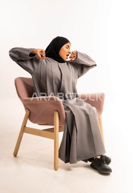 تعابير الخمول و الشعور بالنعاس و التعب, الحاجه الى الراحة و النوم، بورتريه لامرأة عربيه خليجيه سعوديه محجبه ترتدي العباءة تجلس على كرسي مريح ترفع يديها للأعلى بإيماءات التثاؤب، خلفية بيضاء