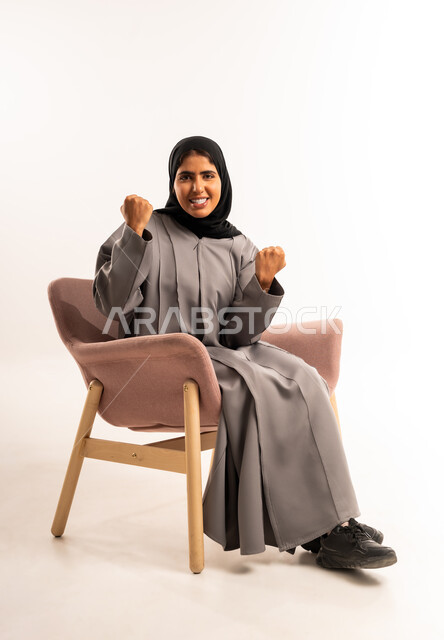 النجاح والفوز وتحقيق الأهداف, القوة والانتصار والتشجيع, بورتريه لشابة عربية خليجية سعودية محجبة مبتسمة ترتدي العباءة تجلس على كرسي مريح ترفع يديها بإيماءات الحماس, النظر إلى الكاميرا بتعابير السرور, خلفية بيضاء
