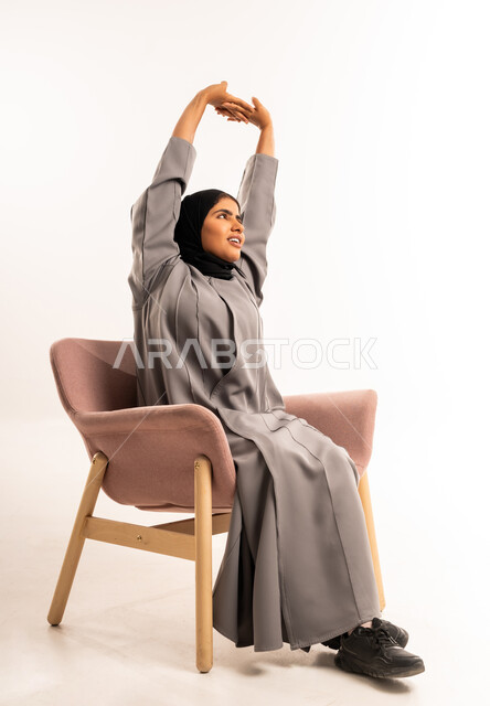 مفهوم الخمول و الكسل, الشعور بالنعاس والحاجه الى الراحة و النوم، بورتريه لامرأة عربيه خليجيه سعوديه محجبه ترتدي العباءة تجلس على كرسي مريح ترفع يديها للأعلى تتمدد بإيماءات التعب، خلفية بيضاء