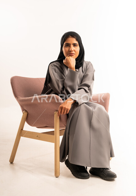 Thinking to find a solution to a problem, absent-mindedness, contemplation and planning for the future, portrait of a veiled Saudi Arabian Gulf woman wearing an abaya sitting on a comfortable chair and looking at the camera with gestures of concentration, facing problems and challenges in practical life, white background