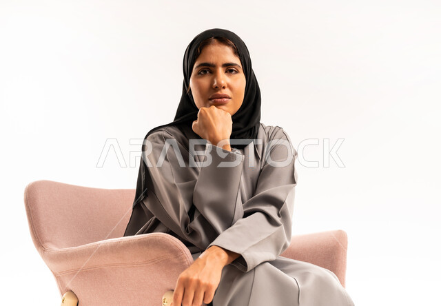 شرود الذهن والتأمل والتخطيط للمستقبل، التفكير لإيجاد حل مشكلة ما، بورتريه لامرأة عربيه خليجيه سعوديه محجبه ترتدي العباءة تجلس على كرسي مريح وتنظر الى الكاميرا بإيماءات التركيز، مواجهة المشكلات والتحديات في الحياة العملية، خلفية بيضاء