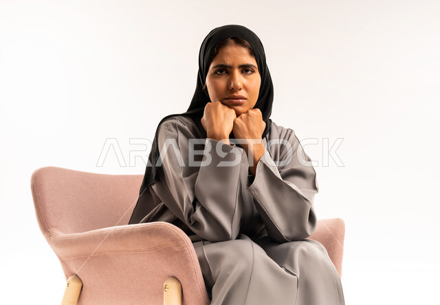 الشعور بالخيبة والانزعاج، إيماءات الحزن والإحباط، الندم على شيء ما، بورتريه مقرب لشابة عربية سعودية خليجية محجبة ترتدي العباءة تجلس على كرسي مريح وتنظر الى الكاميرا بتعابير العبوس, خلفية بيضاء