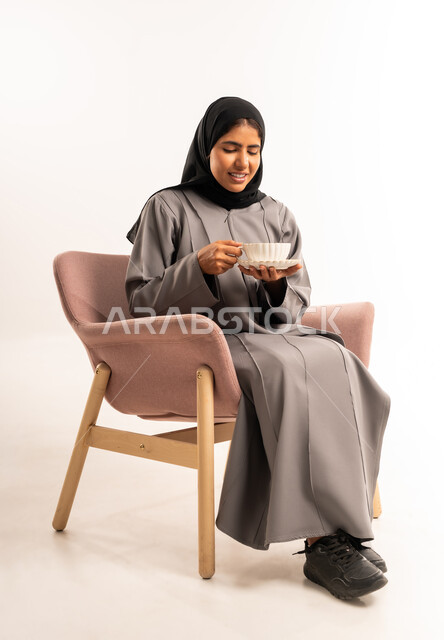 الشعور بالدفء والراحة، الاستعداد لتناول المشروب الساخن، بورتريه لامراه عربية خليجية سعودية مبتسمة محجبة ترتدي العباءة تجلس على كرسي مريح تحمل بيدها فنجان من الشاي بايماءات السرور، خلفية بيضاء