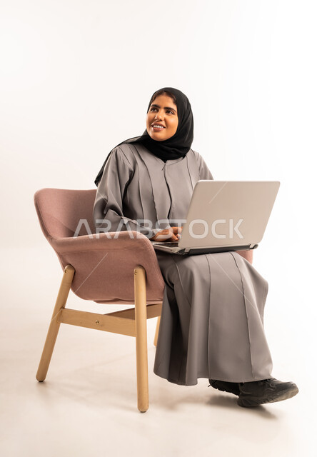 انجاز مهمة العمل عن بعد، مفهوم التسويق عبر الانترنت، بورتريه لامرأة عربيه خليجيه سعوديه محجبه ترتدي العباءة تجلس على كرسي مريح تستخدم اللابتوب المحمول تنظر الى شيء ما بإيماءات السرور، التواصل مع العائلة والاصدقاء, خلفية بيضاء