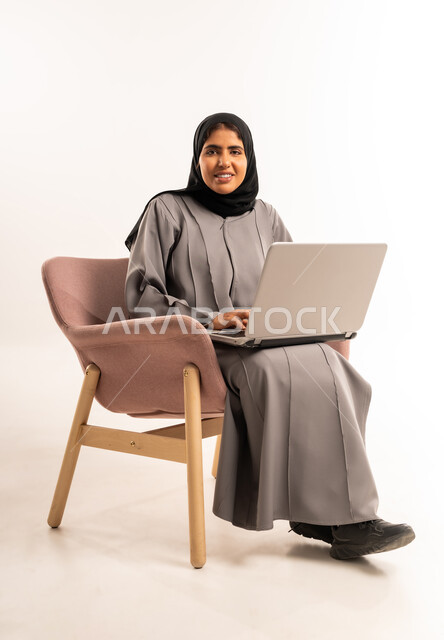 Expressions of cheerfulness and happiness, accomplishing a remote work task, the concept of electronic marketing, a portrait of a smiling Saudi Arabian Gulf woman wearing a hijab, sitting on a comfortable chair, using a laptop, looking at the camera with gestures of self-confidence, white background