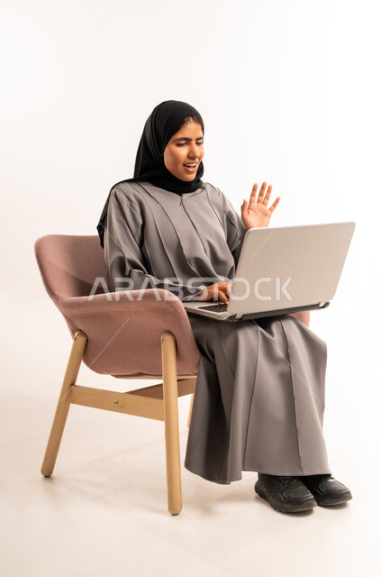 Remote business meeting, video call and live broadcast, portrait of a veiled Saudi Arabian Gulf woman wearing an abaya sitting on a comfortable chair using a laptop raising her hand up with waving and greeting gestures, expressions of cheerfulness and happiness, white background