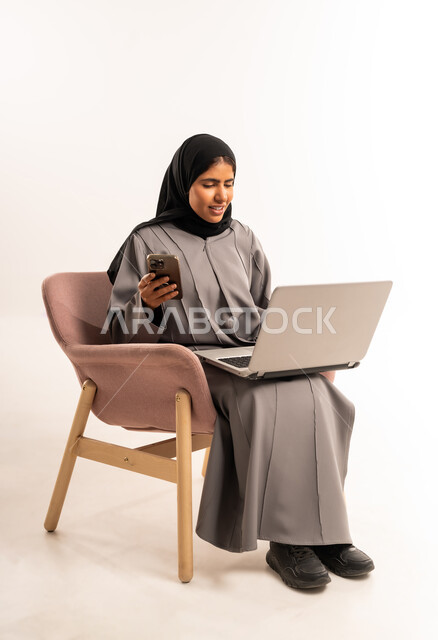 مفهوم التسويق عبر الانترنت، تصفح موقع التواصل الاجتماعي, انجاز مهام العمل عن بعد, بورتريه لامرأة عربية خليجية سعودية مبتسمة محجبة ترتدي العباءة تجلس على كرسي مريح تستخدم الجوال و جهاز اللابتوب المحمول بتعابير السرور, خلفية بيضاء