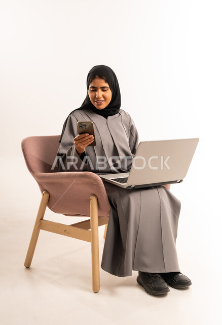 تصفح موقع التواصل الاجتماعي, انجاز مهام العمل عن بعد, مفهوم التسويق عبر الانترنت، بورتريه لامرأة عربية خليجية سعودية مبتسمة محجبة ترتدي العباءة تجلس على كرسي مريح تستخدم الجوال و جهاز اللابتوب المحمول بتعابير السرور, خلفية بيضاء