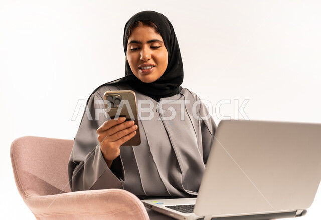 الدردشة مع العائلة والأصدقاء, مفهوم التسويق عبر الانترنت، تصفح موقع التواصل الاجتماعي, بورتريه مقرب لامرأة عربية خليجية سعودية مبتسمة محجبة ترتدي العباءة تستخدم الجوال و جهاز اللابتوب المحمول بتعابير السرور, خلفية بيضاء