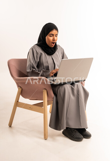 إجراء مكالمة فيديو وبث مباشر، إدارة وتنظيم العمل عن بعد، بورتريه لامرأة عربيه خليجيه سعوديه محجبه ترتدي العباءة تجلس على كرسي مريح تنظر الى جهاز الحاسوب المحمول بإيماءات الدهشة والذهول، الدردشة والتواصل مع الاهل والأصدقاء، خلفية بيضاء