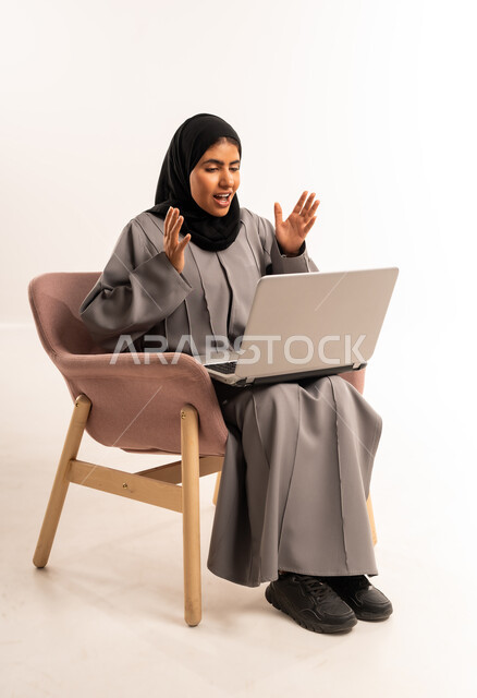 إدارة وتنظيم العمل عن بعد، إجراء مكالمة فيديو، بورتريه لامرأة عربيه خليجيه سعوديه محجبه ترتدي العباءة تجلس على كرسي مريح تنظر الى جهاز الحاسوب المحمول ترفع يديها للاعلى بإيماءات الدهشة والذهول، الدردشة والتواصل مع الاهل والأصدقاء، خلفية بيضاء