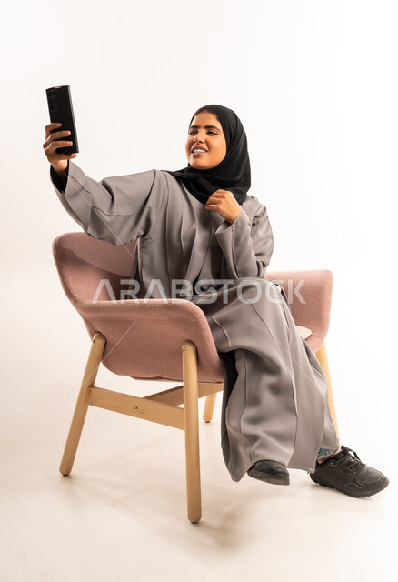 Documenting happy moments, using a modern advanced technical device, portrait of a smiling Saudi Arabian Gulf woman wearing a hijab, sitting on a comfortable chair, holding a mobile phone in her hand, taking a commemorative selfie, expressions of joy and happiness, white background