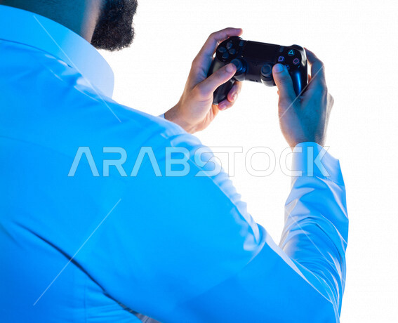 صورة مقربة من الخلف لرجل عربي خليجي سعودي ، يمسك عصا التحكم و يلعب بألعاب الفيديو ، المتعة و التسلية بألعاب الفيديو و ألعاب العالم الافتراضي ، ألعاب البلايستيشن الترفيهية ، تقنيات و ألعاب ، خلفية بيضاء