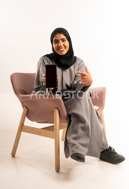 عرض شاشة سوداء فارغة, استخدام جهاز تقني حديث متطور، بورتريه لامرأة عربية خليجية سعودية مبتسمة محجبة ترتدي العباءة تجلس على كرسي مريح تنظر الى الكاميرا بإيماءات السرور تشير بإصبع السبابة إلى الهاتف المحمول, خلفية بيضاء