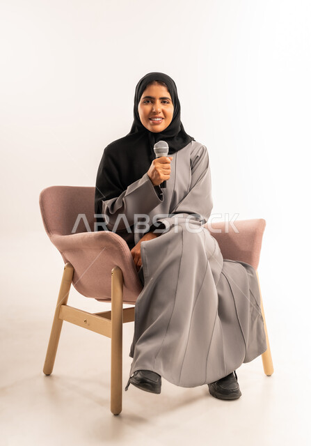 تسجيل مقاطع غنائية، مفهوم التدريب والآداء الصوتي، فن ومهارة تقديم العرض المسرحي, بورتريه لامرأة عربية سعودية خليجية محجبة ترتدي العباءة تجلس على كرسي مريح تمسك بيدها الميكروفون تنظر الى الكاميرا تغني بتعابير الاندماج والتفاعل، خلفية بيضاء