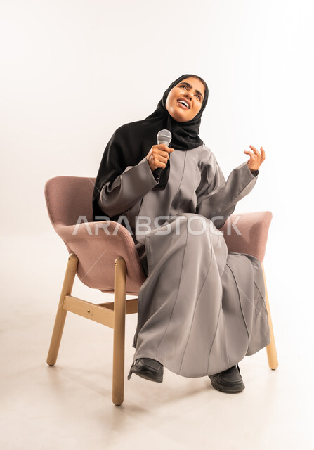 مفهوم التدريب والآداء الصوتي، فن ومهارة تقديم العرض المسرحي, بورتريه لامرأة عربية سعودية خليجية محجبة ترتدي العباءة تجلس على كرسي مريح تمسك بيدها الميكروفون تغني بتعابير الاندماج والتفاعل، تسجيل مقاطع غنائية، خلفية بيضاء