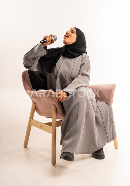 فن ومهارة تقديم العرض المسرحي, بورتريه لامرأة عربية سعودية خليجية محجبة ترتدي العباءة تجلس على كرسي مريح تمسك بيدها الميكروفون تغني بتعابير الاندماج والتفاعل، مفهوم التدريب والآداء الصوتي، تسجيل مقاطع غنائية، خلفية بيضاء
