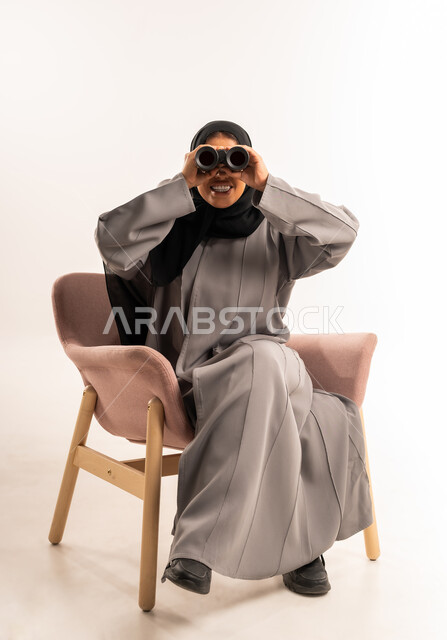مفهوم تقريب الأشياء البعيدة والتخطيط, بورتريه لامرأة عربية سعودية خليجية محجبة ترتدي العباءة تجلس على كرسي مريح تقوم بالنظر من خلال المنظار ثنائي العينين ( التلسكوب )، استكشاف و رؤية مستقبلية للهدف، تعابير الفرح والسرور, خلفية بيضاء