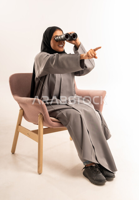 الإشارة بإصبع السبابة الى شيء ما, بورتريه لامرأة عربية سعودية خليجية محجبة ترتدي العباءة تجلس على كرسي مريح تقوم بالنظر من خلال المنظار ثنائي العينين ( التلسكوب )، استكشاف و رؤية مستقبلية للهدف، مفهوم تقريب الأشياء البعيدة والتخطيط, خلفية بيضاء