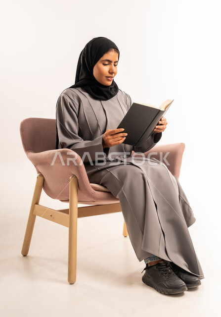 قراءة الرواية والقصة، تعابير التركيز والاستمتاع، تمضية وقت الفراغ بالتعلم والتثقيف الذاتي، بورتريه لامرأة عربية سعودية خليجية محجبة ترتدي العباءة تجلس على كرسي مريح تحمل بيدها كتاب تقرأ بإيماءات الاندماج, خلفية بيضاء
