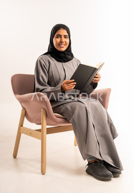 تمضية وقت الفراغ بالتعلم والتثقيف الذاتي، قراءة الرواية والقصة، تعابير التركيز والاستمتاع، بورتريه لامرأة عربية سعودية خليجية محجبة ترتدي العباءة تجلس على كرسي مريح تحمل بيدها كتاب تنظر الى الكاميرا بإيماءات السرور, خلفية بيضاء