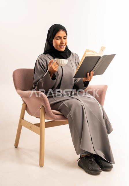 Gestures of concentration and integration, drinking a hot drink, spending free time learning and self-education, reading a novel and story, portrait of a veiled Saudi Arabian Gulf woman wearing an abaya sitting on a comfortable chair holding a book in her hand and drinking tea, white background