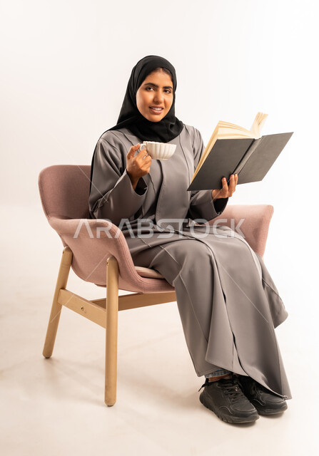 احتساء مشروب ساخن، تمضية وقت الفراغ بالتعلم والتثقيف الذاتي، قراءة الرواية والقصة، بورتريه لامرأة عربية سعودية خليجية محجبة ترتدي العباءة تجلس على كرسي مريح تحمل بيدها كتاب تشرب الشاي تنظر الى الكاميرا بإيماءات السرور, خلفية بيضاء