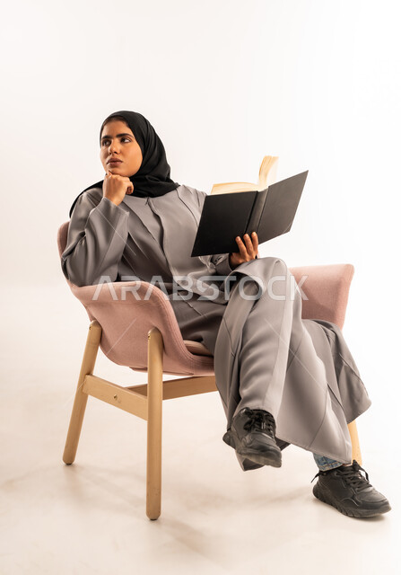 Reading a novel and a story, expressions of focus and enjoyment, spending free time learning and self-education, portrait of a veiled Saudi Arabian Gulf woman wearing an abaya sitting on a comfortable chair holding a book in her hand looking at something with gestures of contemplation, white background