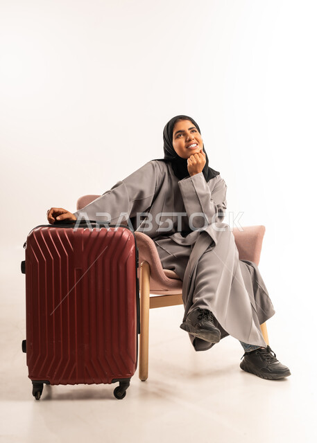 قضاء رحلة صيفية ممتعة, الاستعداد لبدء العطلة خارج البلاد, السياحة في إجازة الصيف, بورتريه لشابة عربية خليجية سعودية محجبة ترتدي العباءة تجلس على كرسي مريح  تضع يدها على حقيبة السفر تنظر إلى الاعلى بإيماءات التأمل والسرور، انتظار موعد الطائرة, خلفية بيضاء