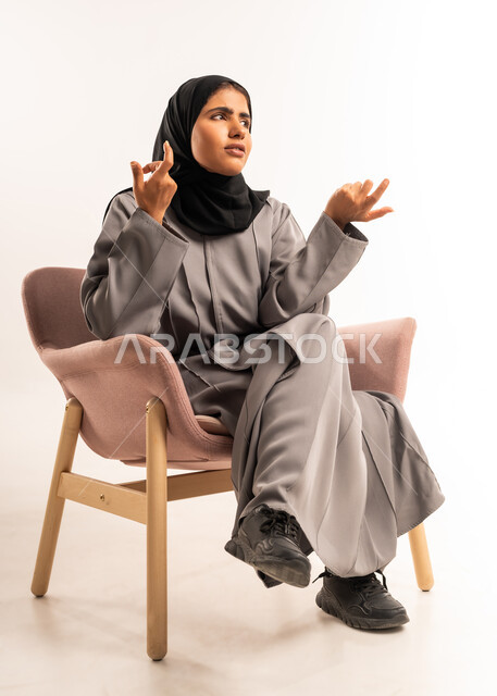 التردد بخصوص أمر معين، الشعور بالجهل وعدم المعرفة، بورتريه مقرب لإمرأة عربية سعودية خليجية محجبة ترتدي العباءة تجلس على كرسي مريح ترفع يديها للأعلى بإيماءات الشعور بالحيرة، خلفية بيضاء