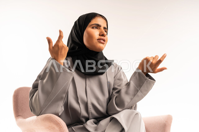 الشعور بالجهل وعدم المعرفة، بورتريه مقرب لإمرأة عربية سعودية خليجية محجبة ترتدي العباءة تجلس على كرسي مريح ترفع يديها للأعلى بإيماءات الشعور بالحيرة، التردد بخصوص أمر معين، خلفية بيضاء