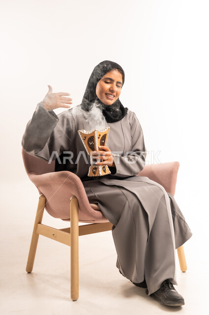 العادات والتقاليد السعودية, استخدام خشب العود بالمناسبة و العيد, التطيب بالعود والبخور, بورتريه لشابة عربية خليجية سعودية محجبة ترتدي العباءة تجلس على كرسي مريح تقوم بالتعطر من حامل البخور, خلفية بيضاء