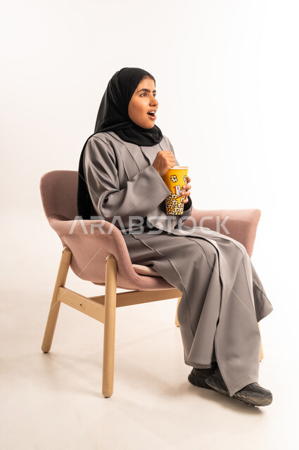 Entertainment by watching series and movies, spending fun and entertaining time at home, gestures of surprise and shock, portrait of a veiled Saudi Arabian Gulf woman wearing an abaya sitting on a comfortable chair holding a cup full of popcorn with expressions of surprise and astonishment, white background