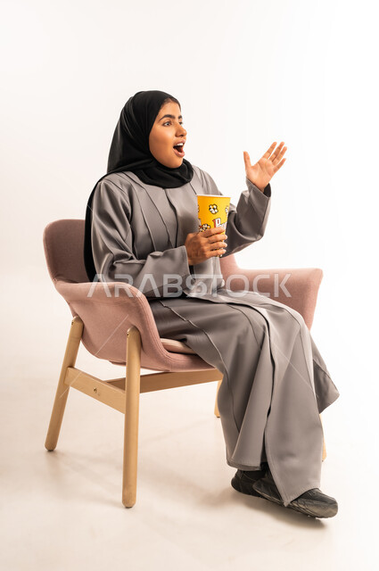قضاء وقت ممتع و ترفيهي في المنزل، ايماءات المفاجأة و الصدمة، التسلية بمتابعة المسلسل و الفيلم، بورتريه لإمرأة عربية خليجية سعودية محجبة ترتدي العباءة تجلس على كرسي مريح تحمل بيدها كوب مليء بالفوشار بتعابير الدهشة و التعجب، خلفية بيضاء