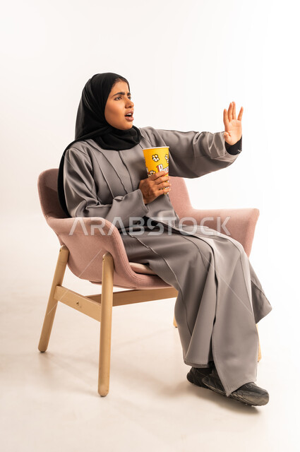 ايماءات المفاجأة و الصدمة، التسلية بمتابعة المسلسل و الفيلم، بورتريه لإمرأة عربية خليجية سعودية محجبة ترتدي العباءة تجلس على كرسي مريح تحمل بيدها كوب مليء بالفوشار بتعابير الدهشة و التعجب، قضاء وقت ممتع و ترفيهي في المنزل، خلفية بيضاء