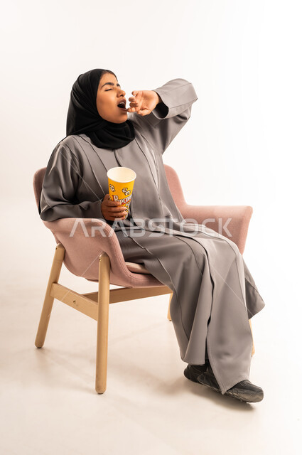 التسلية بمتابعة المسلسل و الفيلم، بورتريه لإمرأة عربية خليجية سعودية محجبة ترتدي العباءة تجلس على كرسي مريح تحمل بيدها وعاء مليء بالفوشار بتعابير النعاس، قضاء وقت ممتع و ترفيهي في المنزل، ايماءات التثاؤب و الحاجة الى النوم، خلفية بيضاء