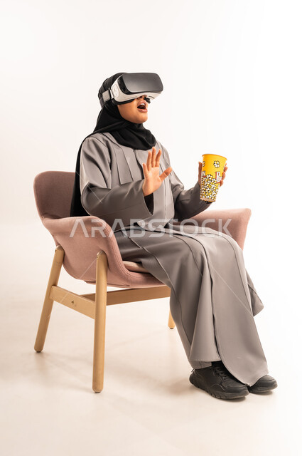 رفع اليد للاعلى بتعابير المفاجأة، التسلية بمشاهدة الفيلم و المسلسل, التفاعل والاندماج مع الواقع الافتراضي، بورتريه لشابة عربية خليجية سعودية محجبة تجلس على كرسي مريح ترتدي العباءة ونظارة في آر ثلاثية الأبعاد تحمل بيدها كوب مليء بالفوشار, خلفية بيضاء