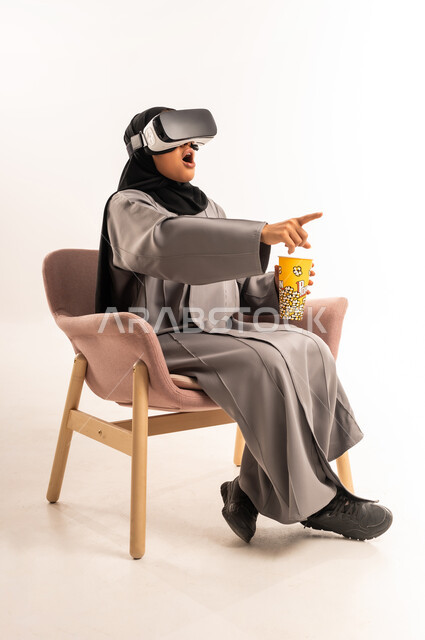 رفع اصبع السبابة و الاشارة الى شيء ما بتعابير المفاجأة، التفاعل مع الواقع الافتراضي، التسلية بمشاهدة الفيلم والمسلسل, بورتريه لشابة عربية خليجية سعودية محجبة تجلس على كرسي مريح ترتدي العباءة ونظارة في آر ثلاثية الأبعاد تحمل بيدها كوب مليء بالفوشار, خلفية بيضاء